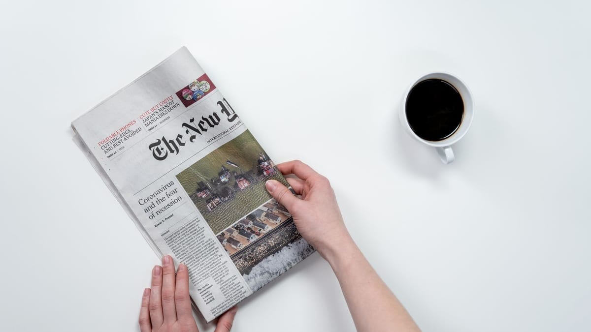 Flat lay of hands holding The New York Times newspaper with coffee on a white table.