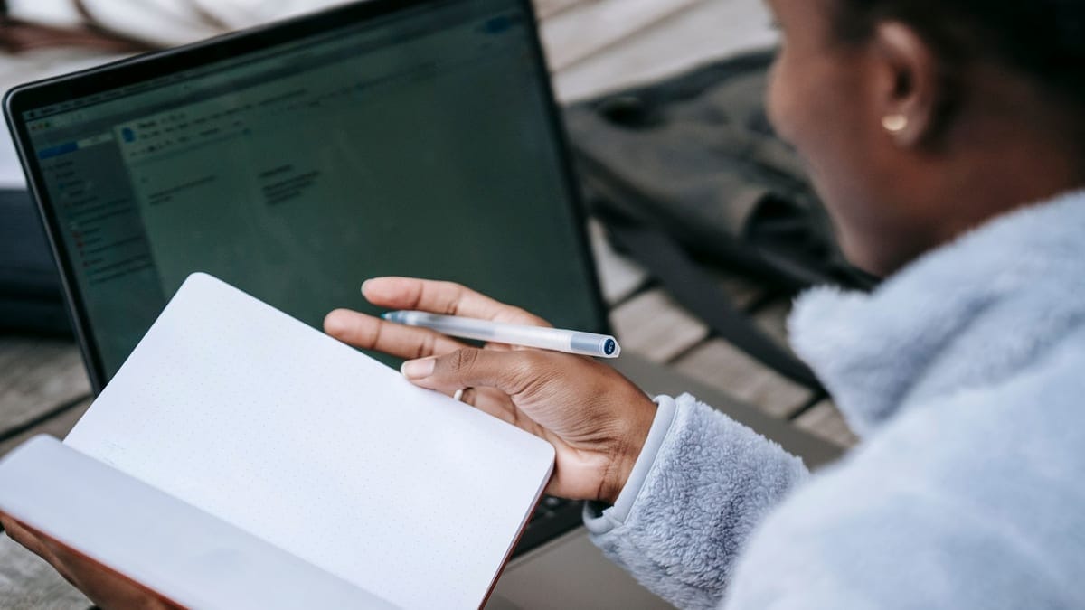High angle of crop unrecognizable African American female student taking notes in notebook while working on research using laptop in campus