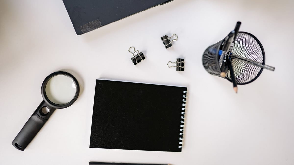 Flat lay of a workspace featuring a laptop, black notebook, magnifying glass, and office supplies on a white surface.