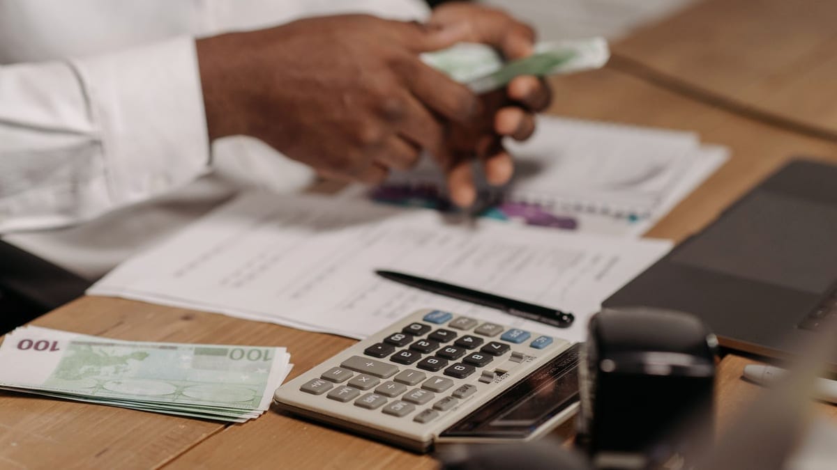 Hands counting euro bills on a wooden desk with calculator, financial documents, and laptop nearby.