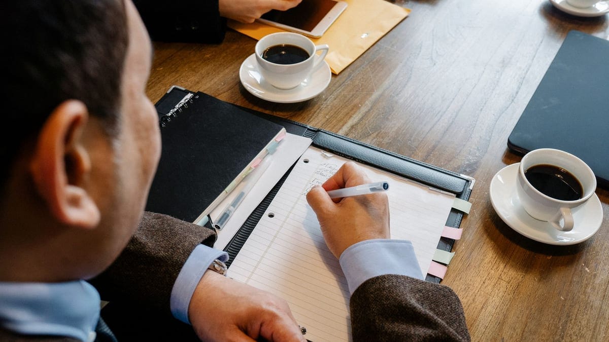 Crop unrecognizable man in formal clothes sitting at table with cup of coffee and writing notes in notepad during business meeting