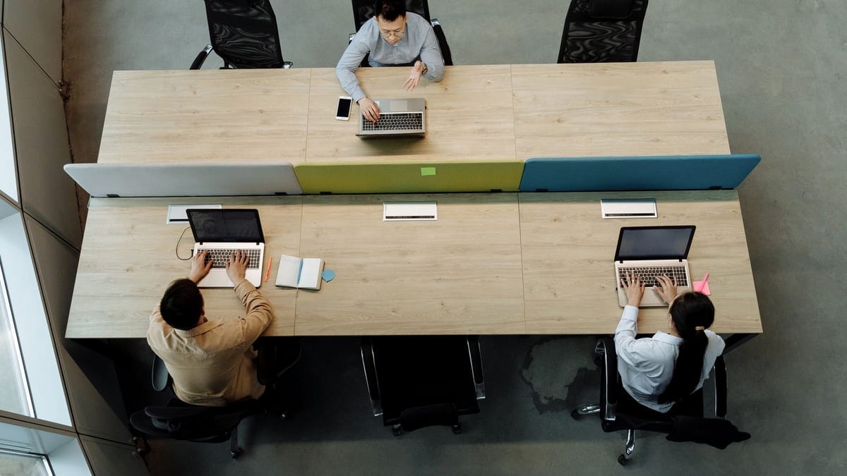 Three colleagues working at a shared office desk, using laptops in a modern workspace.